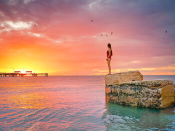 A woman in a red bathing suits stands atop an old piece of a fort on a coastline, offering perspective of one of the most beautiful sunsets in Florida.