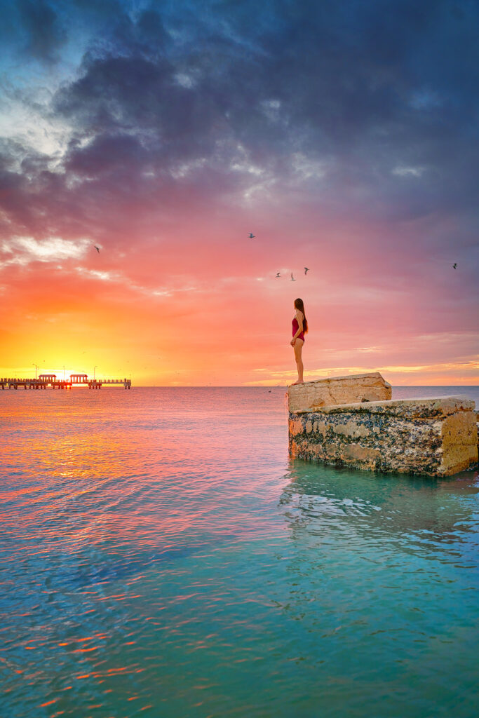 A woman in a red bathing suit stands on the edge of an abandoned fort piece, and birds fly above her, as she look at the sun setting. 