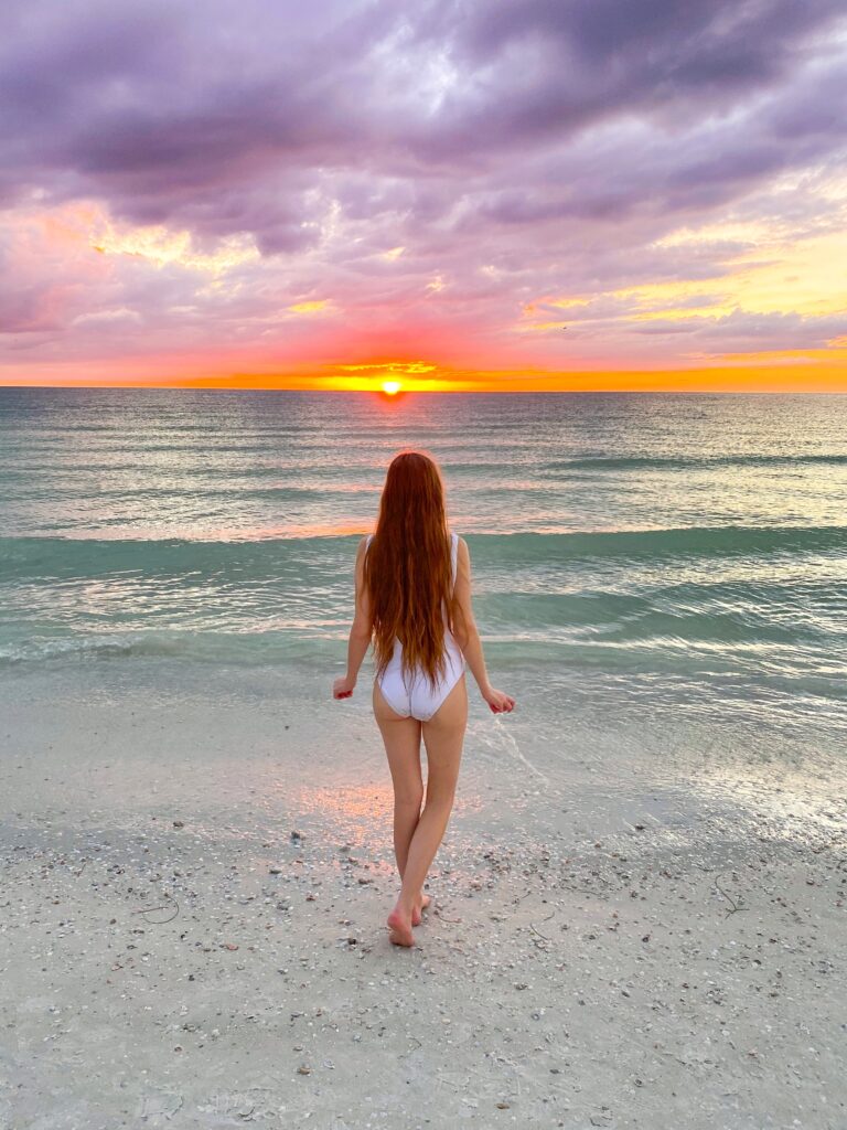 A woman in a white-bathing suit looks over the clear waves and white sand, right at the purple and orange sunset. 