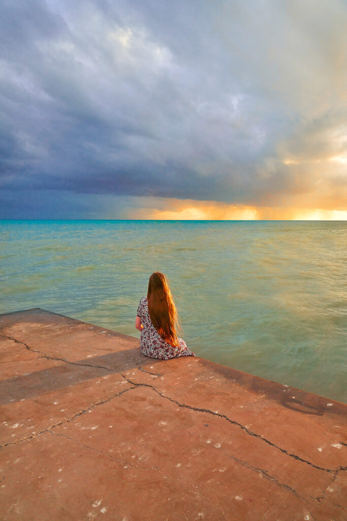 A woman sits on the edge of a sea wall, looking out over the water at a sunset that is yellow and blue.