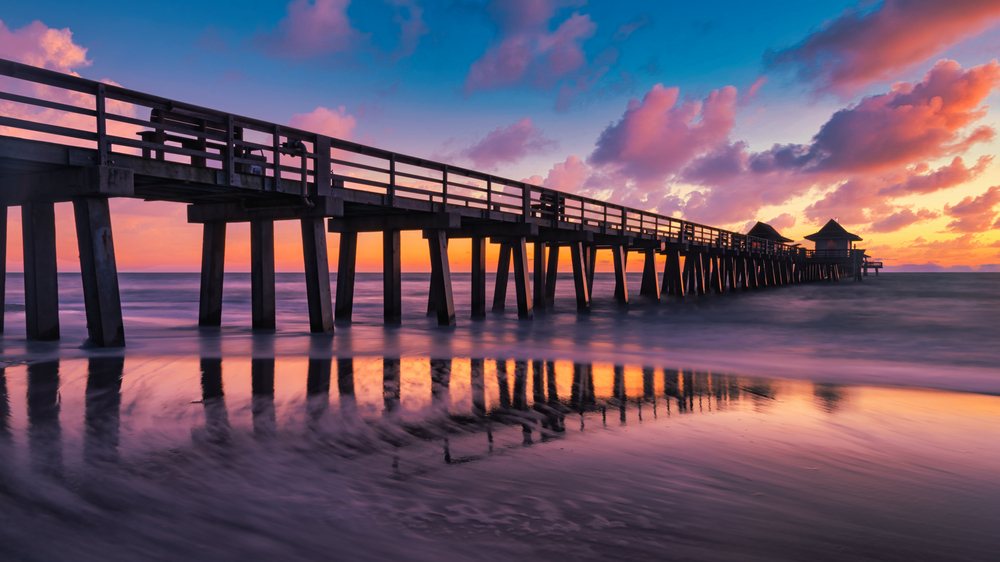 The sunset reflects in the calm water and soft sand in this photo: oranges, purples and blues surround the clouds above the pier. 