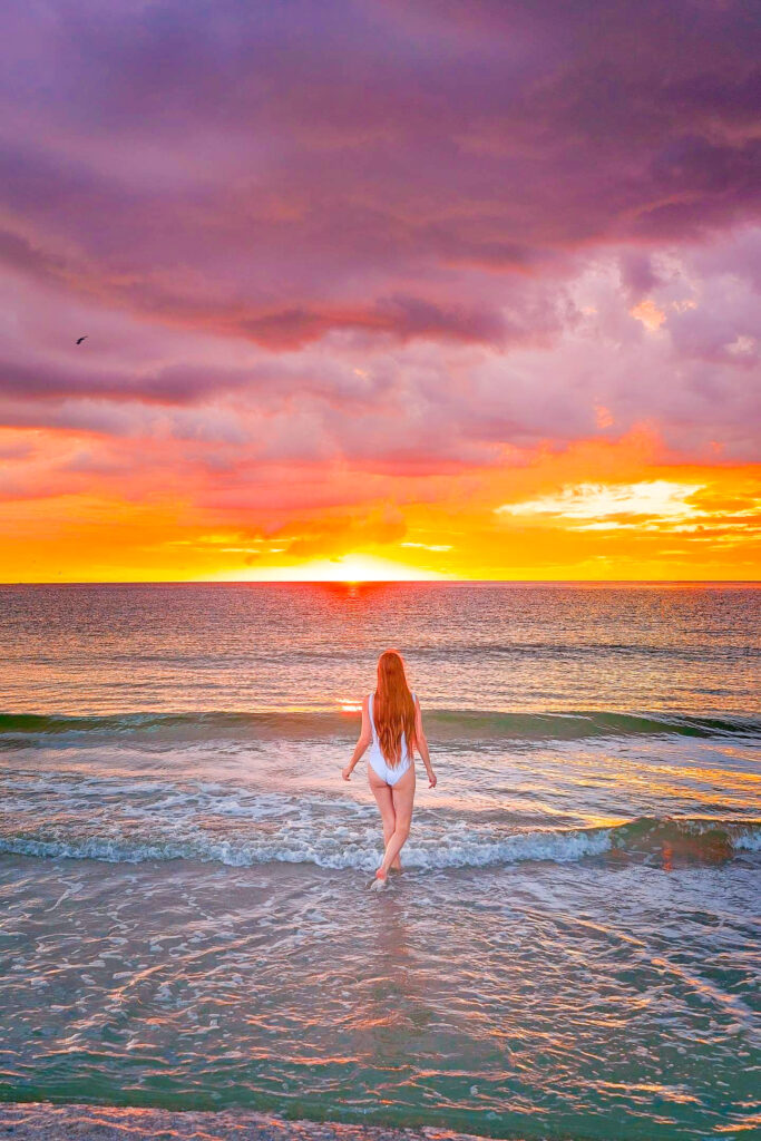 A woman in a white-one-piece looks out at the bright orange sunset while waves crash at her feet. 