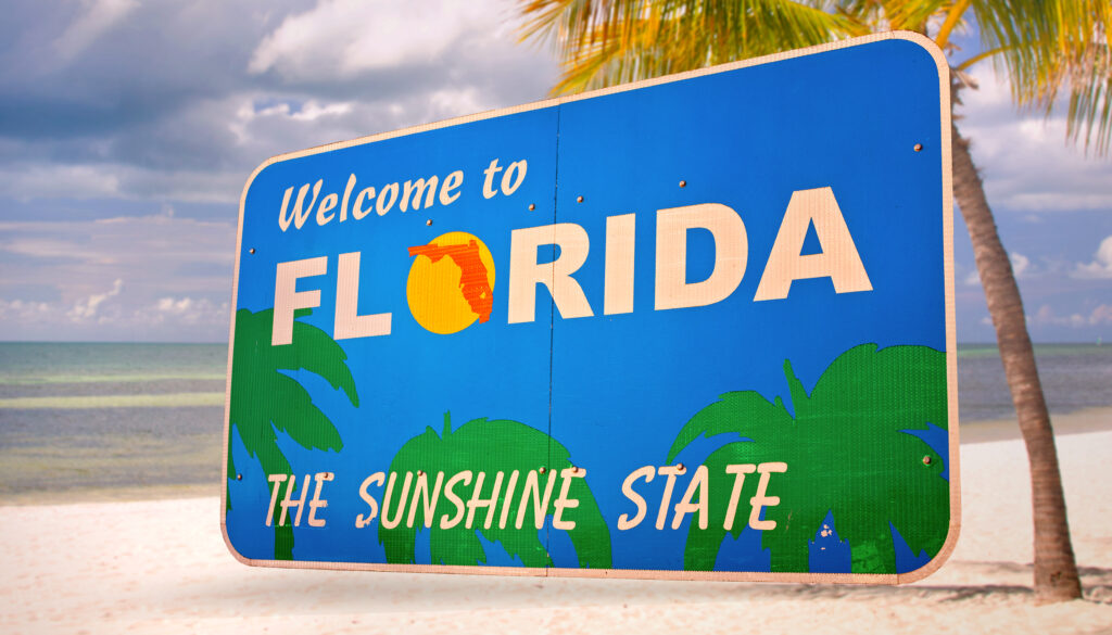 The welcome to Florida sign that reads "the sunshine state" sits in front of a palm tree and on the beach! 