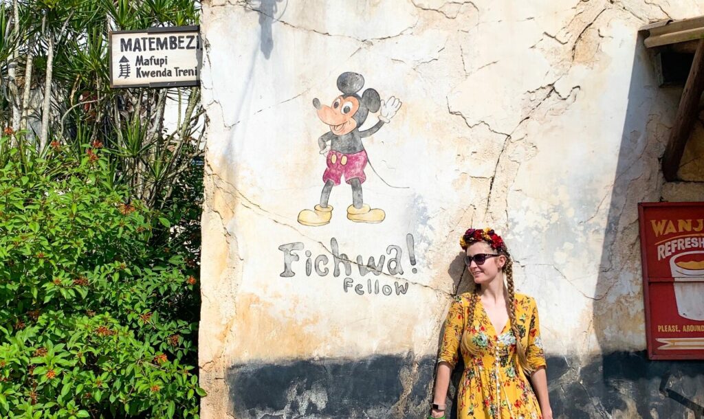 A women in a long yellow dress and braids stands in front of a hidden Mickey at Animal Kingdom, taking a rest. 