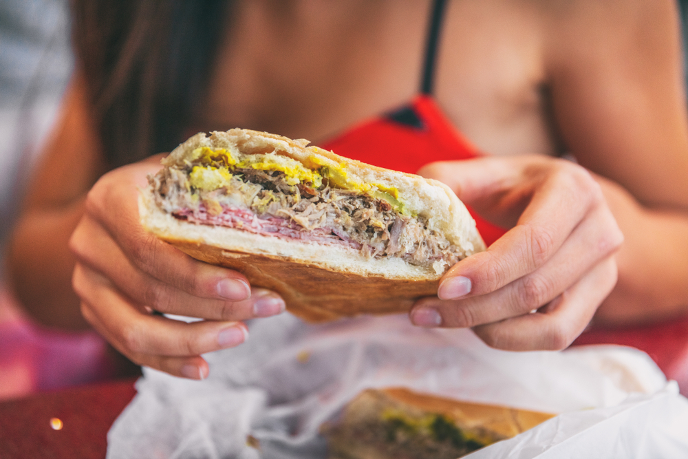 A pair of hands holds a half of a Cuban sandwich, filled to the brim with mustard, pork, pickles and more: one major mistake in Florida is to avoid trying local cuisine like this! 