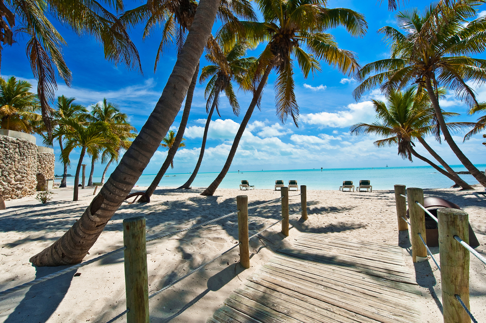 A footbridge connects the mainland to the beach at the keys: blue waters are seen in the distance and palms hover over the bridge itself. 