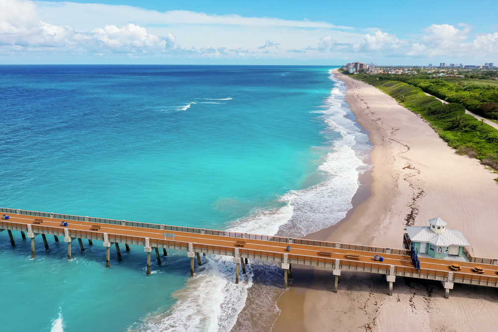 This overhead view of the pier at Juno beach shows off the stunning blue waters, the tan sands, and the greenery behind the pier. 