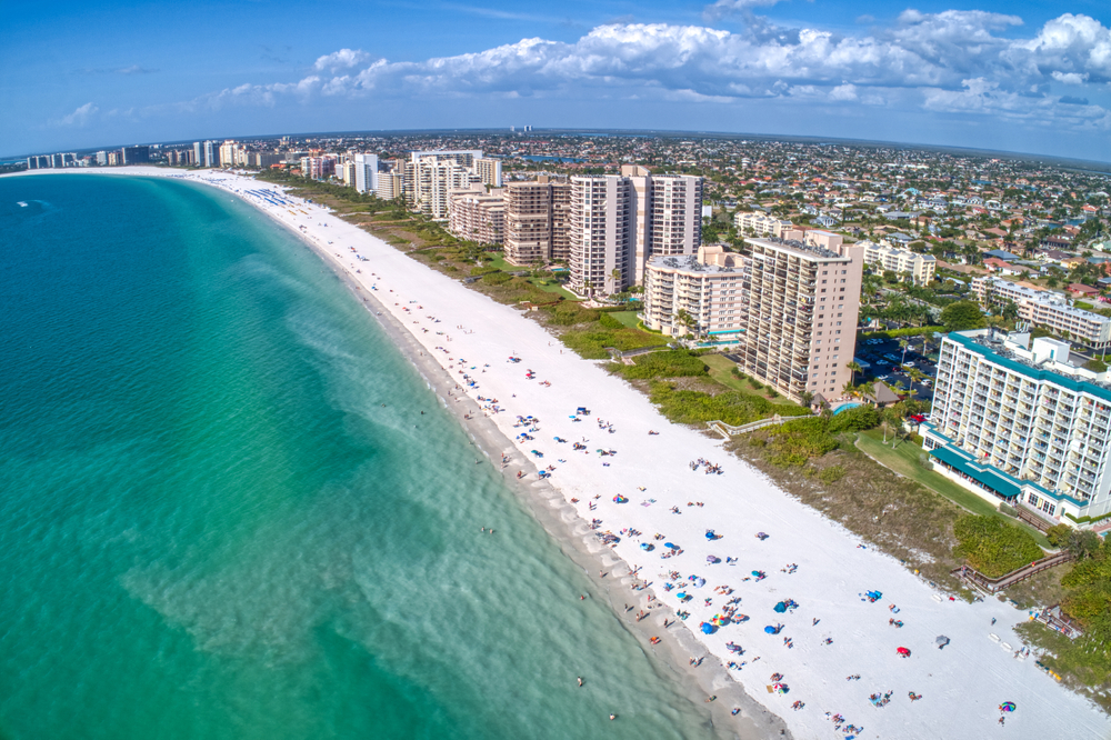 An arial shot of Marco Island shows tall resorts and hotels that tower near the coast: the white sands are dotted with people and the blue water is beautiful. 