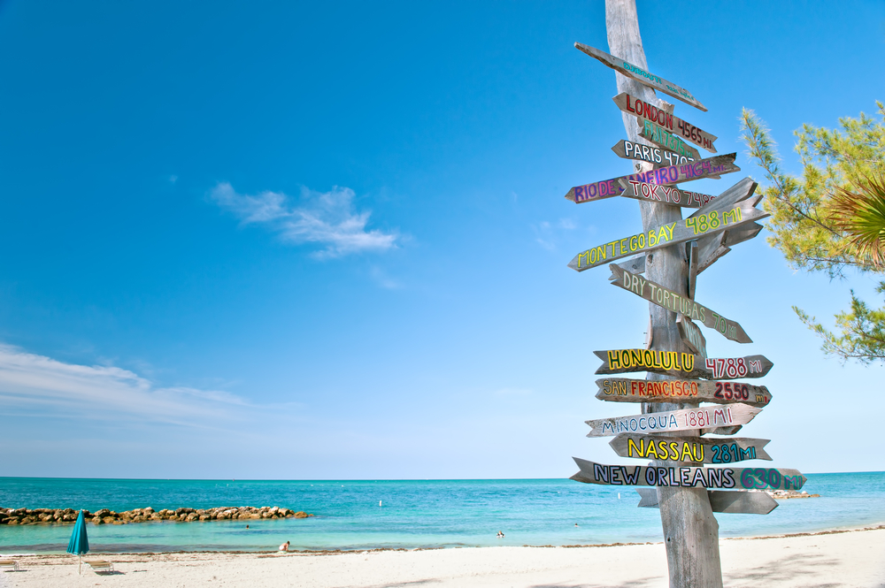 A sign at the keys labels all the near by locations, pointing out the distance of famous cities and beaches as it overlooks the bluest beach water and white sands with a rocky jetty at one of the warmest beaches in Florida. 