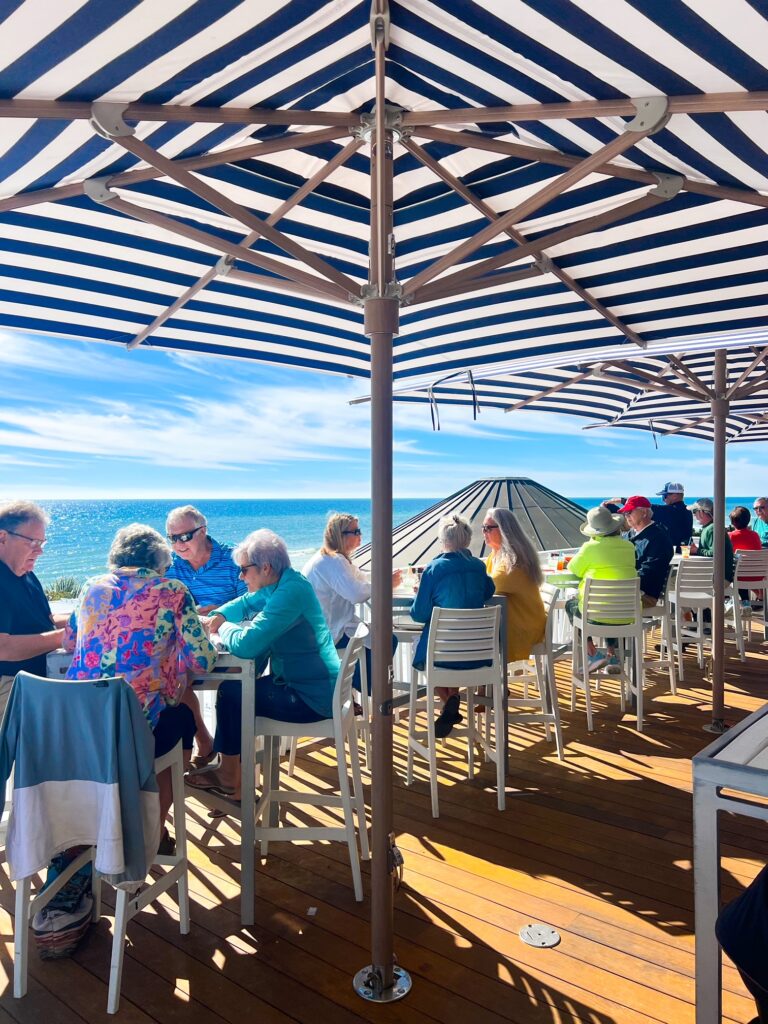 a group of people dining under blue and white umbrellas on a rooftop deck of bud an Allys overlooking the ocean