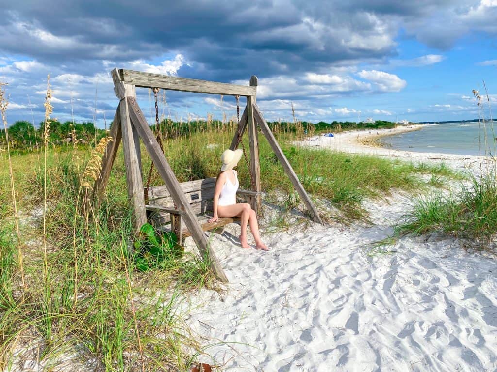 a girl in white swim suit and hat sitting on a wooden swing  on a sandy beach path with sea grass overlooking the ocean