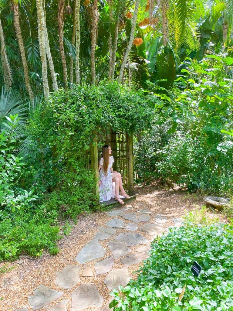 a girl in summer dress sitting on a park bench surrounded by lush Florida foliage in a garden