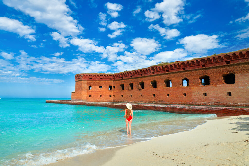 a woman standing on the water edge in a red bathing suit and a white sun hat, a brick wall of a fort is standing in front of the woman