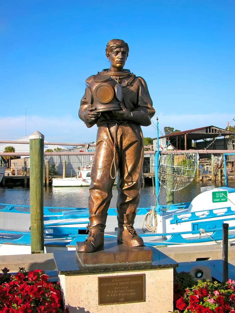 a statue stands near the water, boats are behind it and there are flowers on both side of the statue 