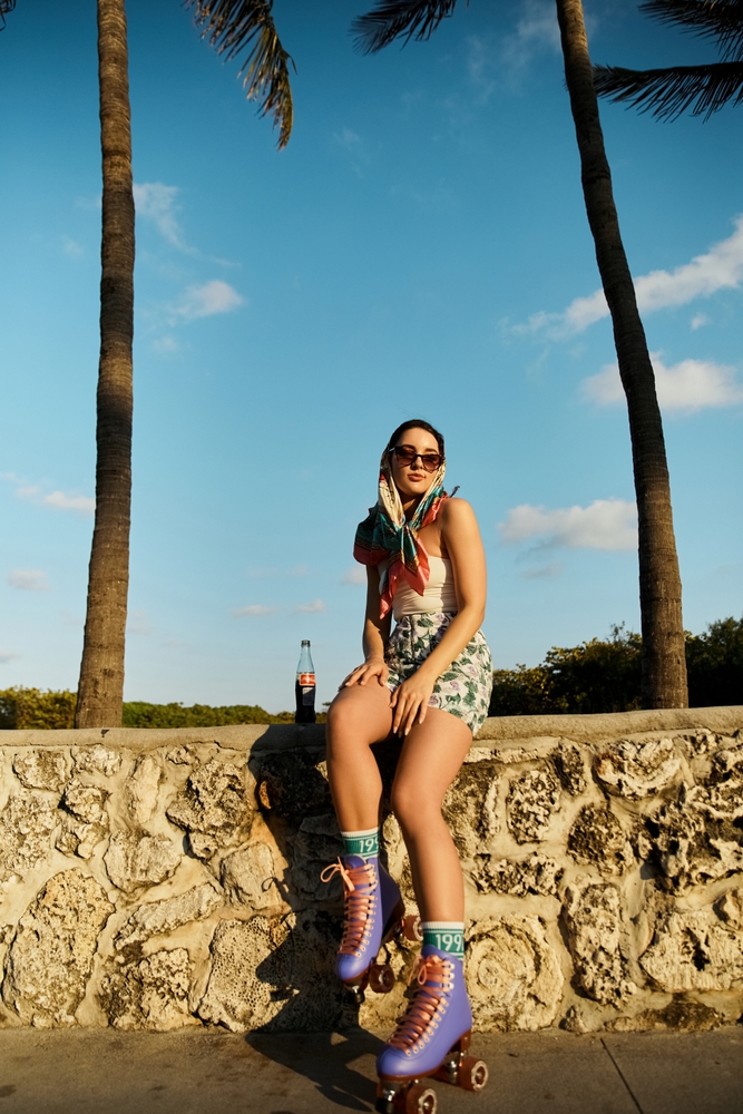 A woman roller skating along a sunlit boardwalk, wearing a scarf and sunglasses, with palm trees behind her and a soft drink bottle beside her on a stone wall.