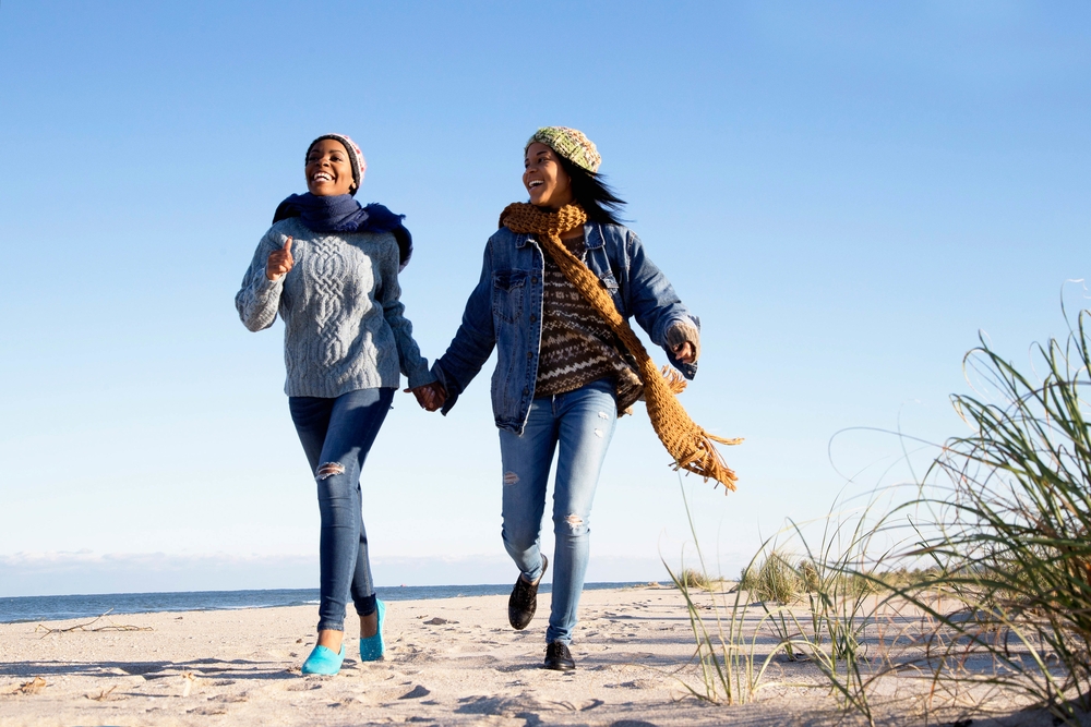 Two women in cozy sweaters and scarves run hand-in-hand along a sandy beach, smiling as they enjoy the crisp air under a bright blue sky.