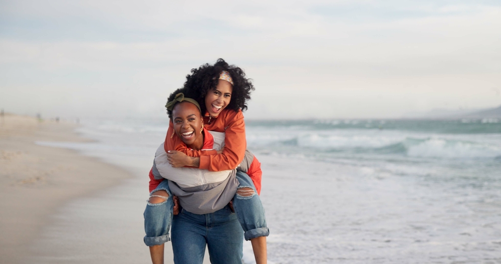 A woman gives a playful piggyback ride to another woman on a beach. They are both laughing, wearing casual jackets and jeans, with the ocean waves in the background.