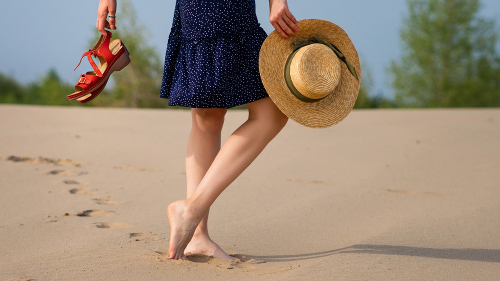 A woman in a polka-dot dress walks barefoot on the sand, holding her shoes in one hand and a wide-brimmed straw hat in the other. She leaves a trail of footprints behind her