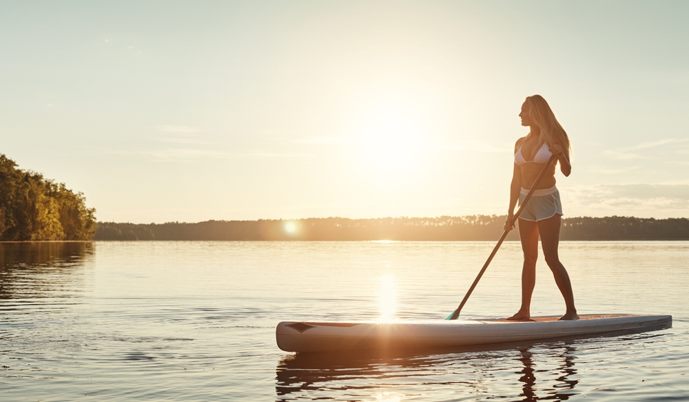 A woman in a bikini top and shorts stands on a paddleboard, enjoying the peaceful lake at sunset, with the golden light reflecting off the water.