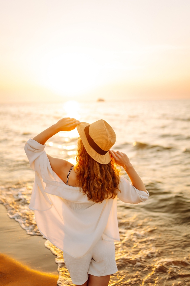 A woman dressed in a light, flowy outfit stands on the shore, holding her straw hat while watching the sun set over the ocean, the golden light reflecting on the water.