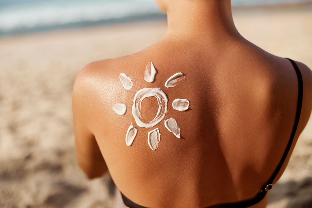 A close-up of a woman's back with sunscreen applied in the shape of a sun, sitting on a sandy beach with the ocean in the background.