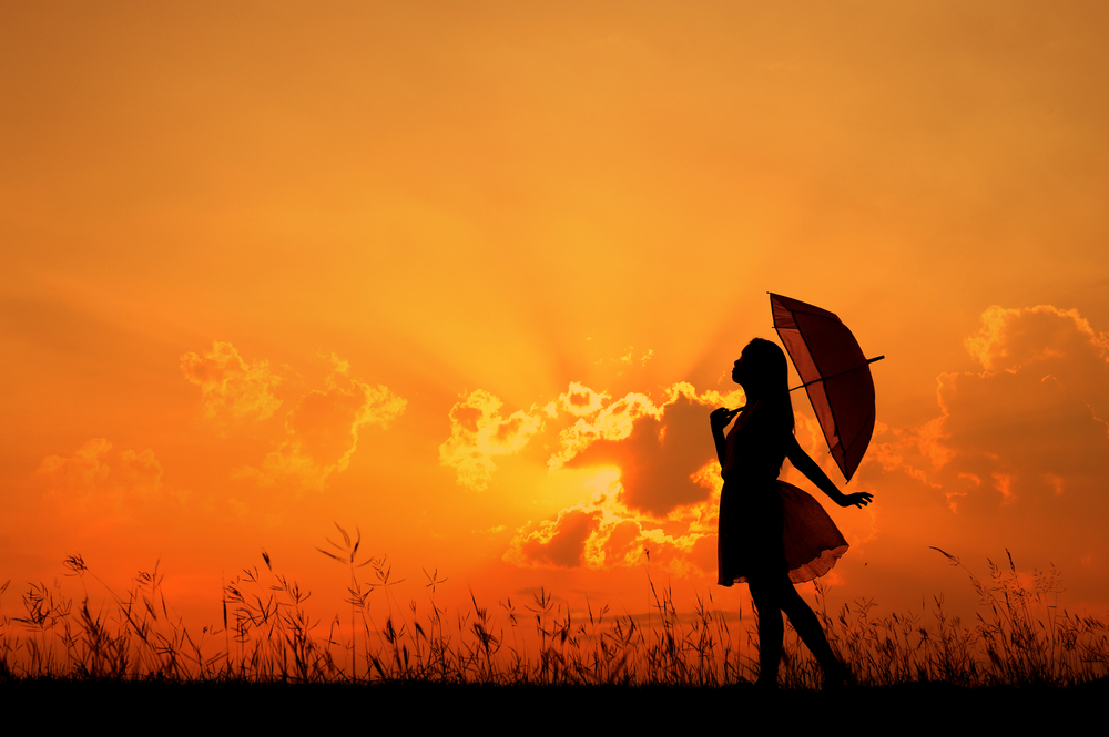 Silhouette of a woman holding an umbrella against a vibrant orange sky during sunset, standing in a field with tall grass.