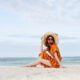 woman sitting on the beach wearing an orange dress and beige sunhat