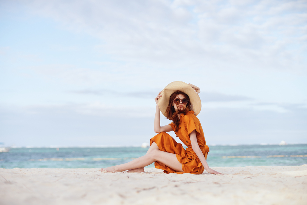 woman sitting on the beach wearing an orange dress and beige sunhat