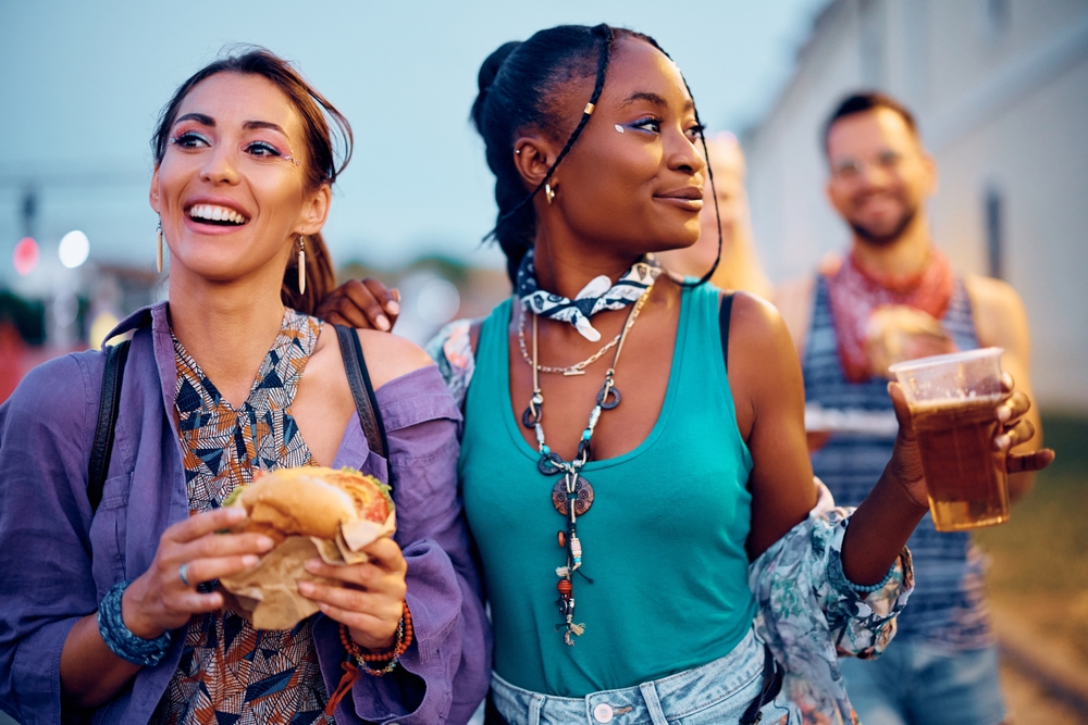 two women standing next to each other at a festival, the one on the left is holding a sandwich and the one of the right is a holding a plastic cup of beer 