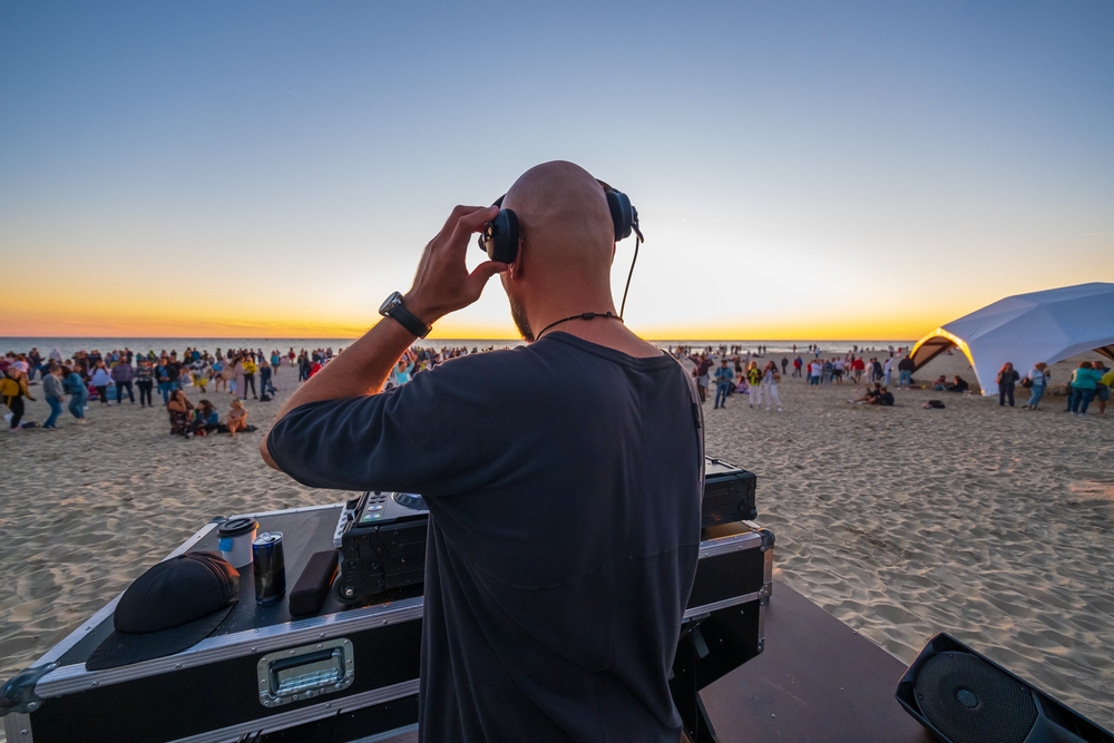 a man is DJing on the beach, he is wearing headphones and there is a crowd in front of him on the sand 