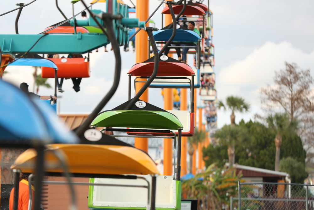 a skylift at florida state fair, there are many seats and there are people sitting in most of the seats, palm trees are in the back 