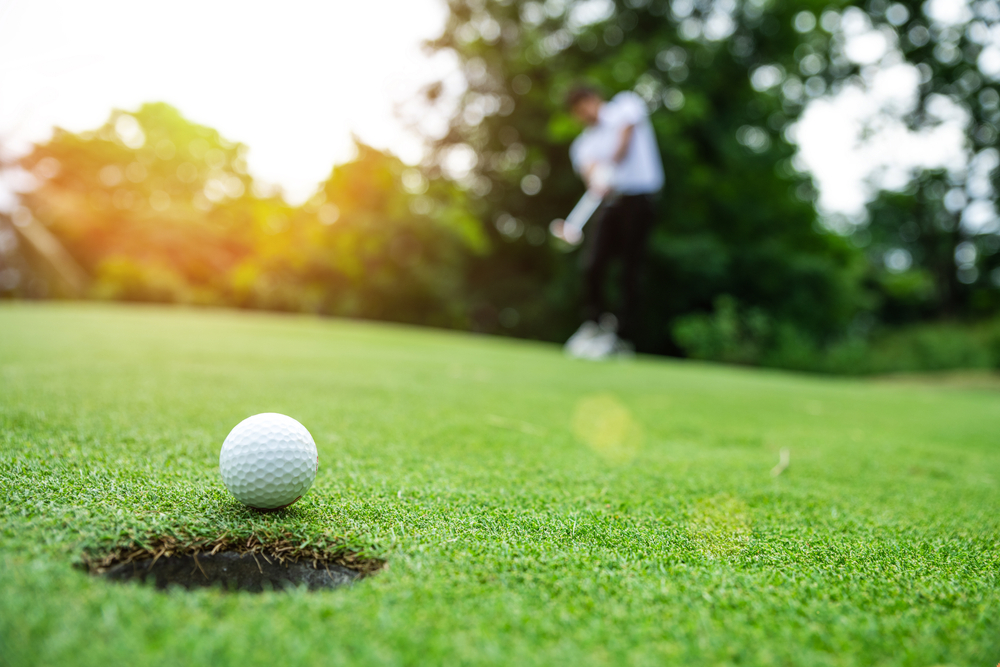 close up of a golf ball near a hole on the golf course, a ma is swinging at the ball in the background