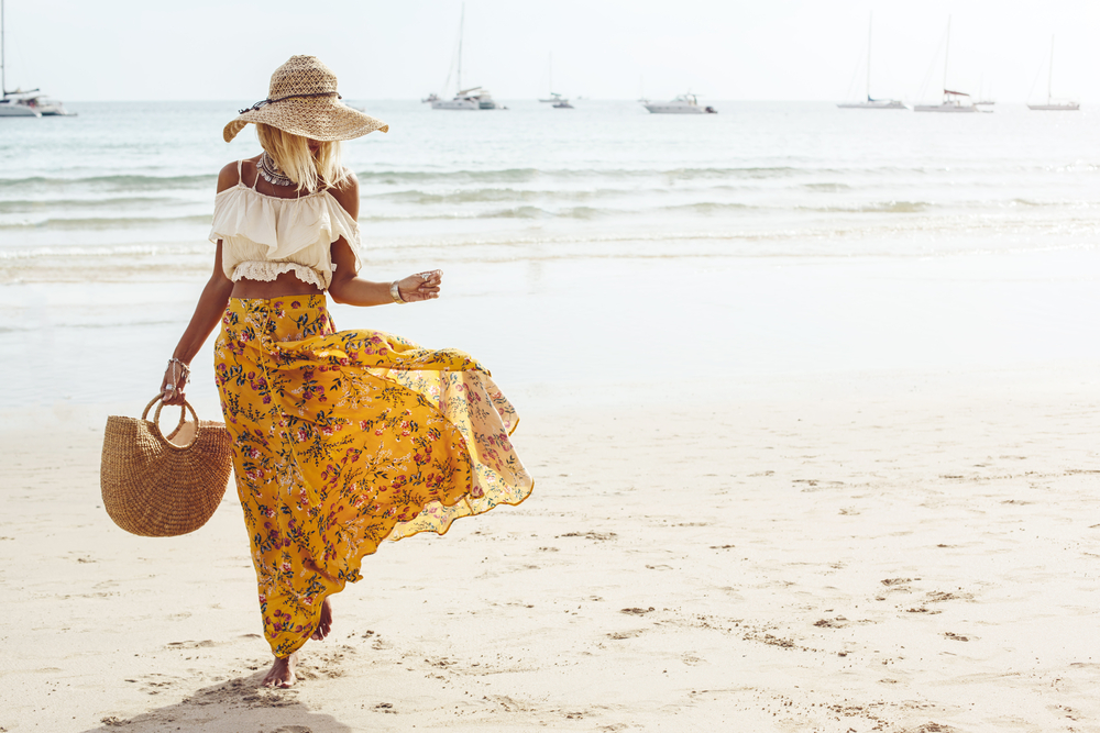 a woman in maxi skirt and crop top with hat and straw bag walking on the beach for what to wear in Florida in summer