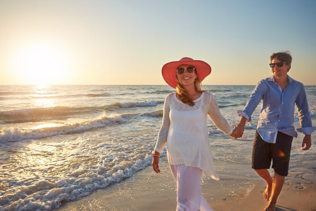 couple walk hand in hand on beach she is wearing a hat. Article is about What to Wear in Florida .   