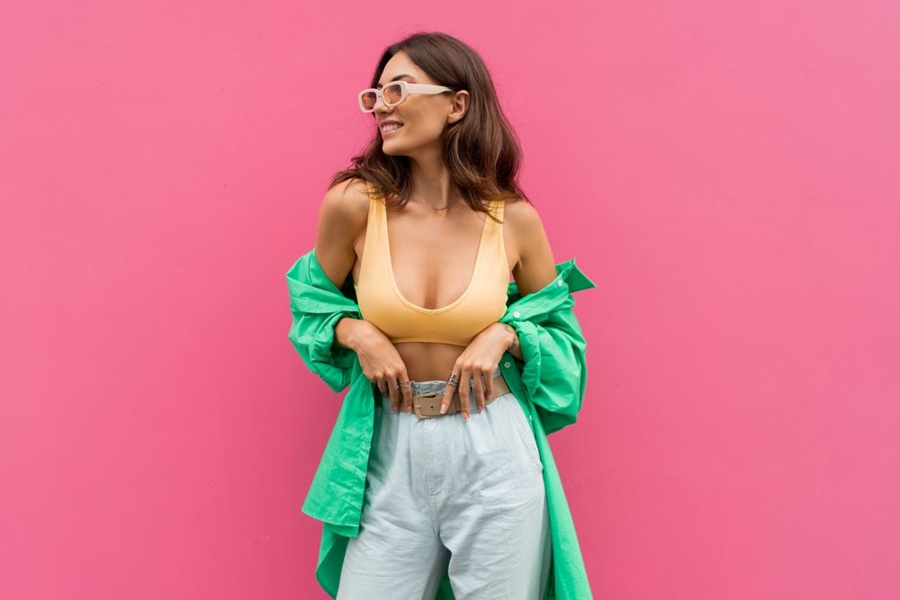 a girl standing on a pink wall with a green unbuttoned shirt, trousers and a sports bra top