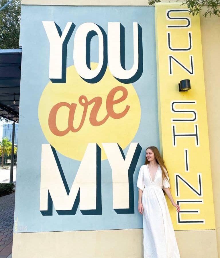 a girl in white dress standing infront of a you are my sunshine mural