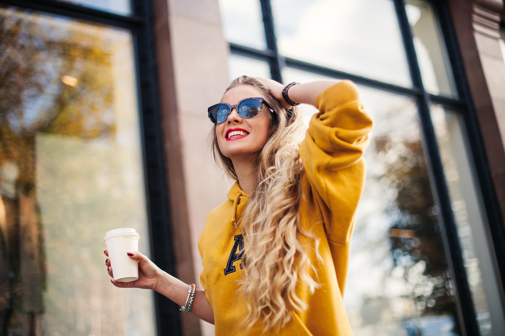 young beautiful blonde drinking coffee walking around the city.mustard sweetshot.,urban backpack , bright red lips. Article is about what to wear in Florida in winter 