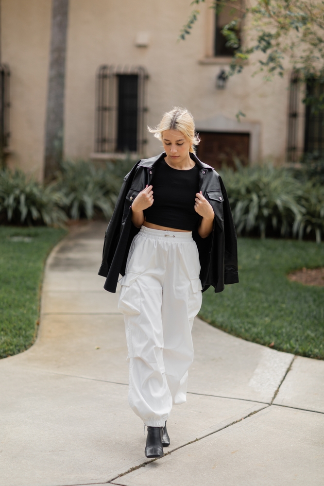 full length of young woman in white cargo pants and leather shirt jacket walking near house in Miami. 