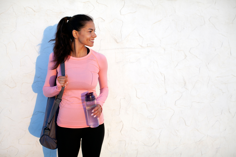 A women in athletic wear leans against a wall, water bottle in hand, comfortable and cool, not worrying about what to wear in Florida in spring. 