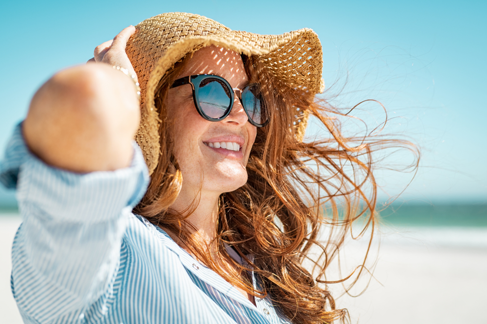 A woman holds onto her big straw hat and sunglasses as the sun and wind dip across her while she stands on the beach, not worrying about what to wear in Florida in spring. 