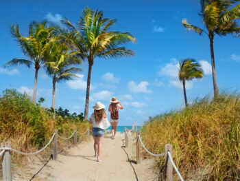 Two girls walk down the sunny path in Florida in spring, in jeans and tank tops and sun hats, not worrying about what to wear in Florida in spring beneath the blue skies and palm trees.