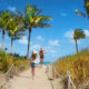 Two girls walk down the sunny path in Florida in spring, in jeans and tank tops and sun hats, not worrying about what to wear in Florida in spring beneath the blue skies and palm trees.