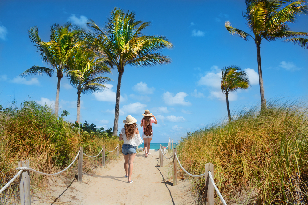 Two girls walk down the sunny path in Florida in spring, in jeans and tank tops and sun hats, not worrying about what to wear in Florida in spring beneath the blue skies and palm trees.