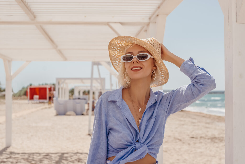 A woman poses on the beach with white sunglasses, a straw hat, and a button down linen shirt hat is blue and flowy. 