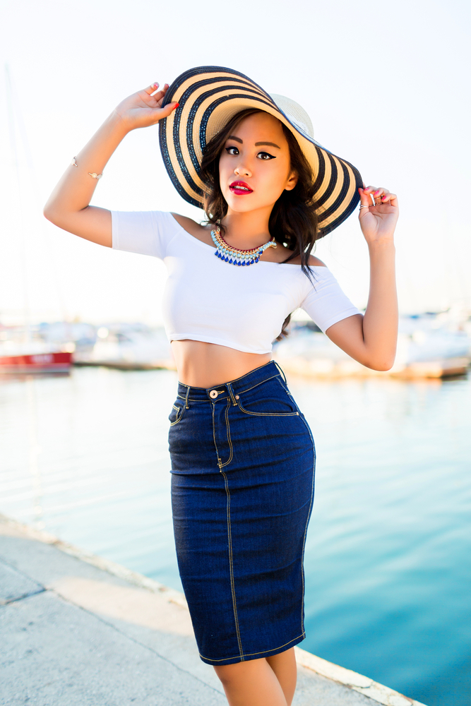 A woman poses near the water in a white crop top and a high waisted denim skirt with statement jewelry and a striped sun hat, channeling a more chic look when considering what to wear in Key West. 