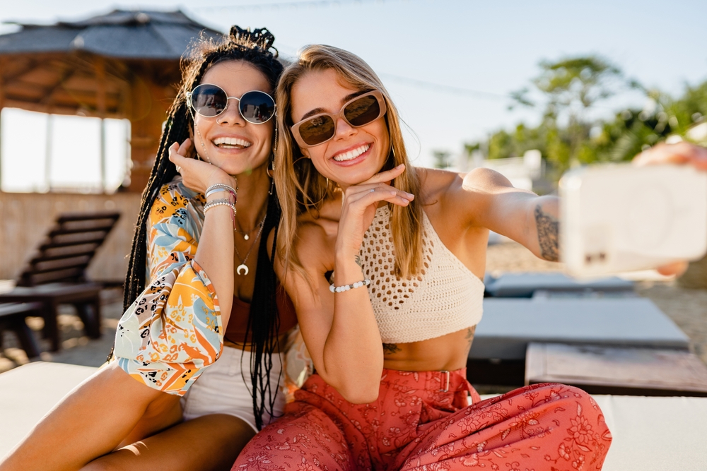 Two friends sit on the beach, both posing in beach attire with flowy pants and shirts, sunglasses and earthy tones. They smile for a selfie, confident in their decisions on what to wear in Key West. 