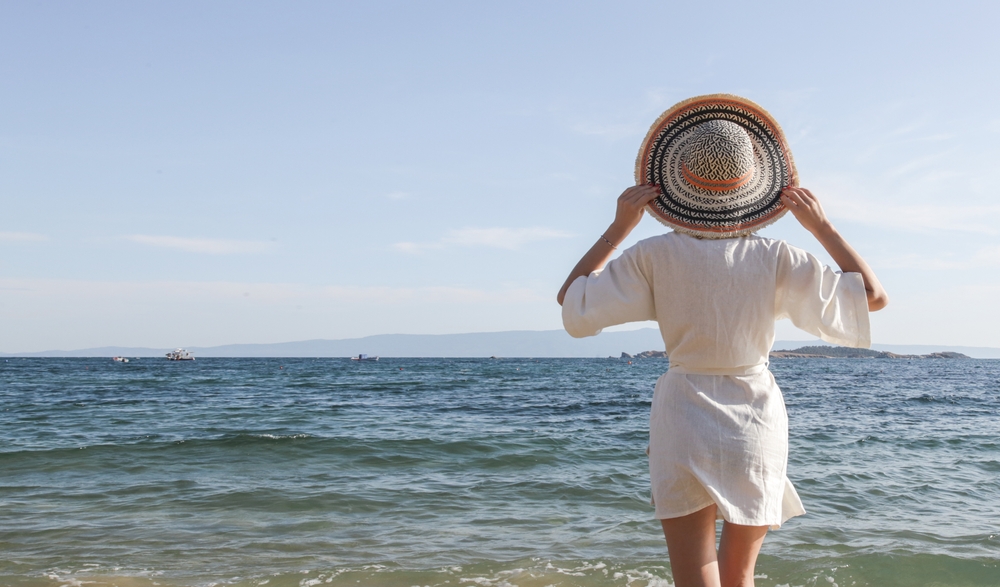 A woman overlooks the ocean, holding onto her hat, in a linen beach cover up. 