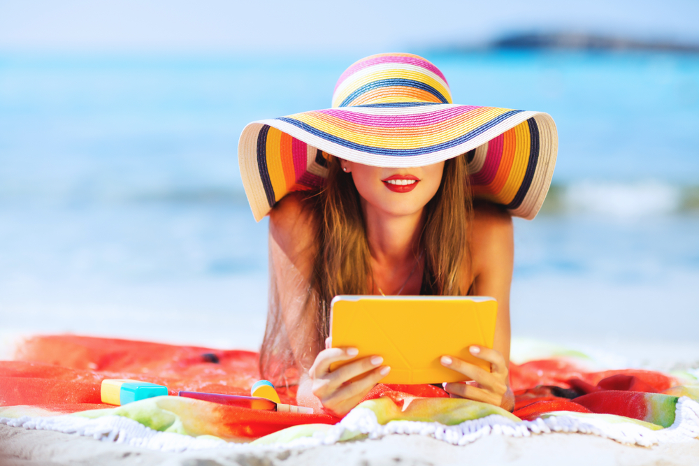 A women sits on the beach with a wide brimmed hat and a kindle: she protects her self from the sun, which is great for what to wear in Miami. 