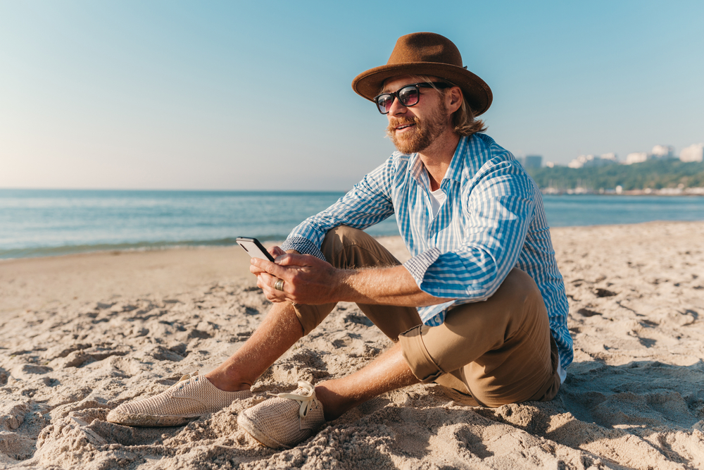 A man sits on the beach in tan pants and a relaxed button down, hat and sunglasses, which is perfect for beach vibes for what to wear in Miami. 