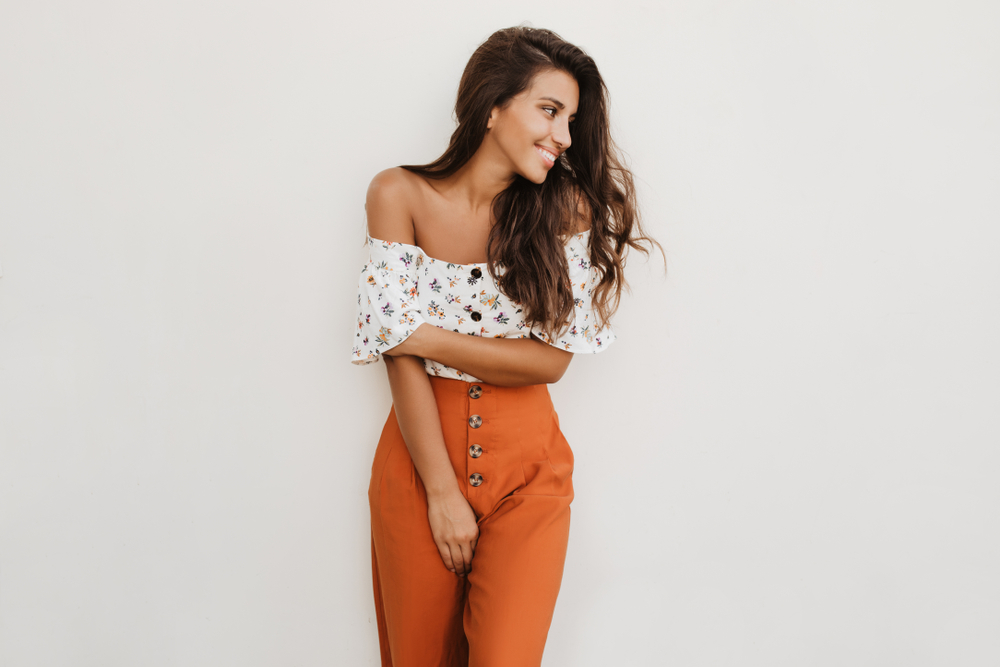 A woman poses in in high waisted orange pants and a romantic blouse with floral print: she smiles and looks off to the right. 